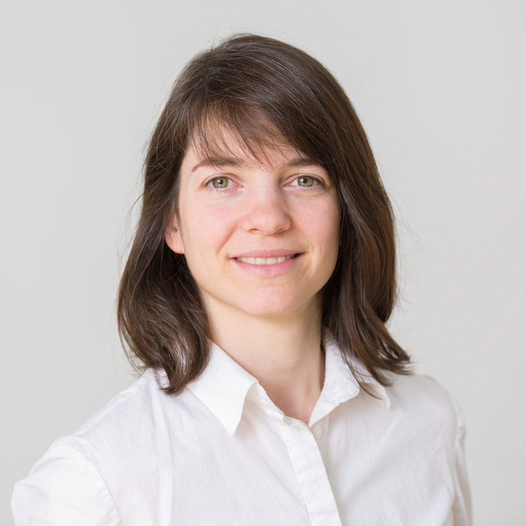 Eine Frau mit Schulter-langem Haar trägt ein weißes Hemd und lächelt freundlich in die Kamera.