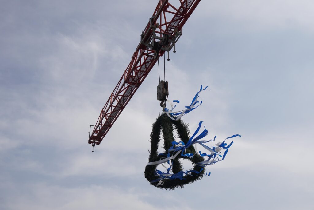 An einem Kran schwingender Nachbau einer Glocke aus grünen Nadeln und flatternden Bändern.