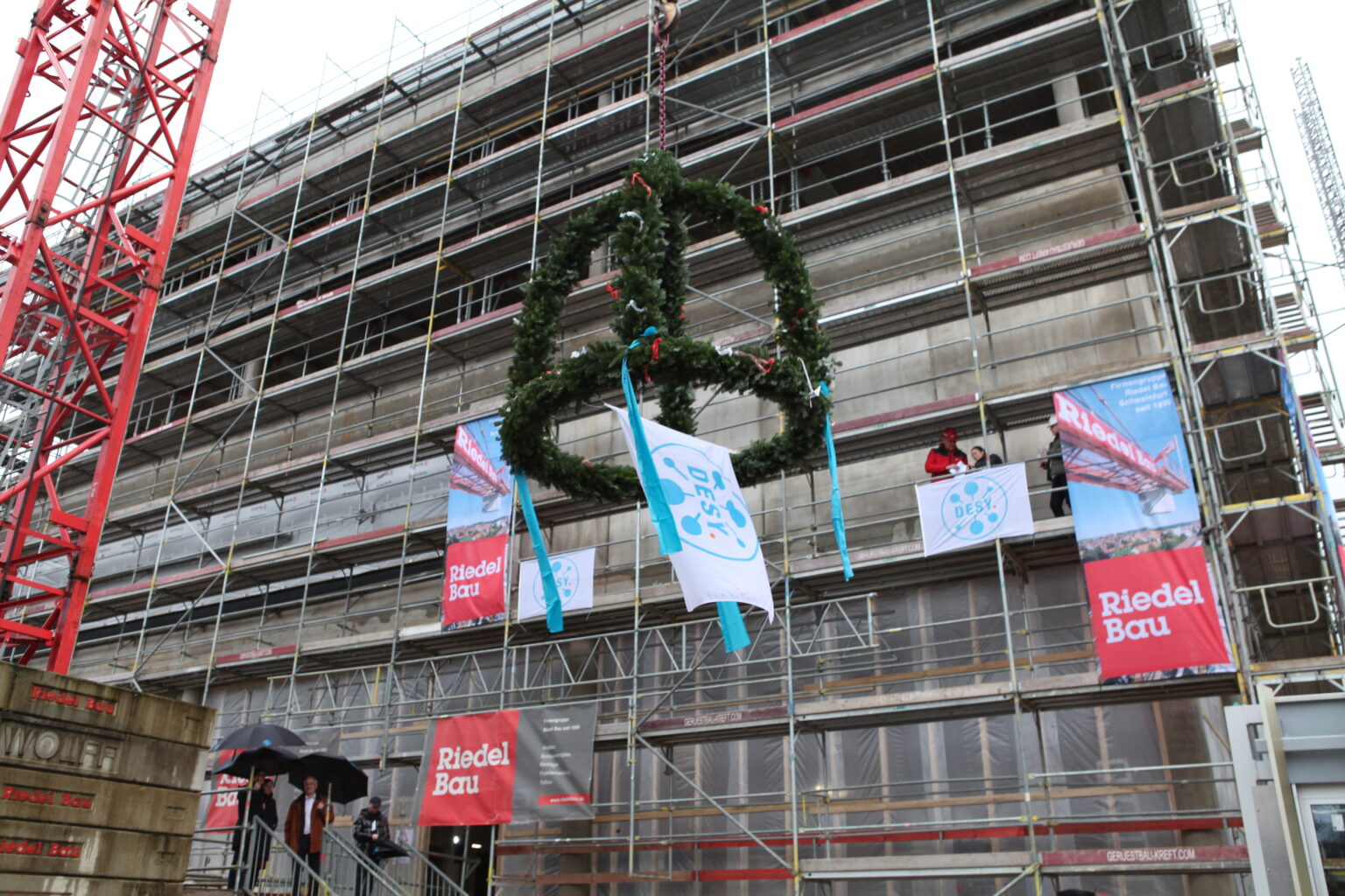 A building under construction is surrounded by scaffolding to which several "Riedel Bau" banners are attached. In the foreground, a topping-out wreath with green branches and blue ribbons is being hoisted by a crane. A flag with the "DESY" logo is attached to the wreath.