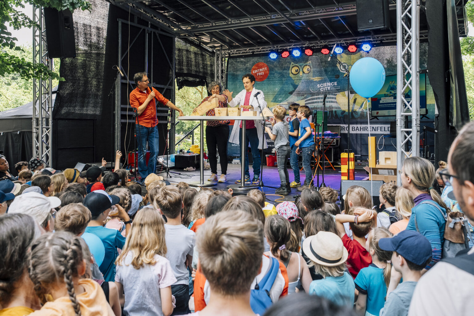 Eine Gruppe von Menschen, hauptsächlich Kinder, versammelt sich vor einer Bühne, auf der Erwachsene spielerisch mit anderen Kindern interagieren.
