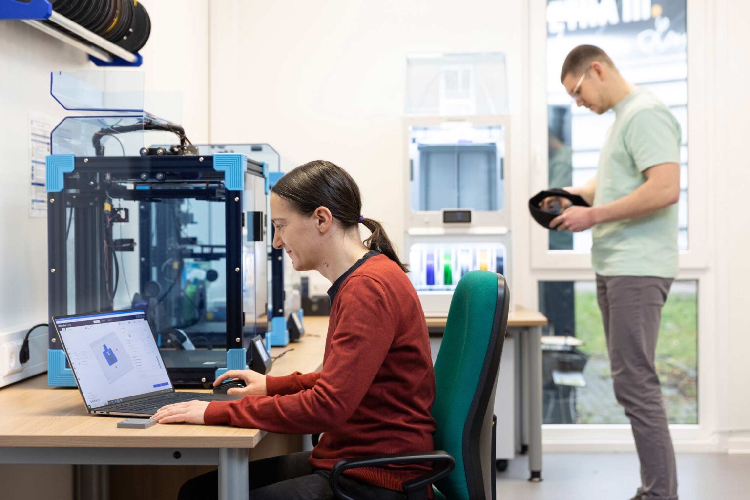 Photo in DESY's Makerspace, where one person is creating a model for 3D printing on the computer. At the second workstation, another person collects material for 3D printing.