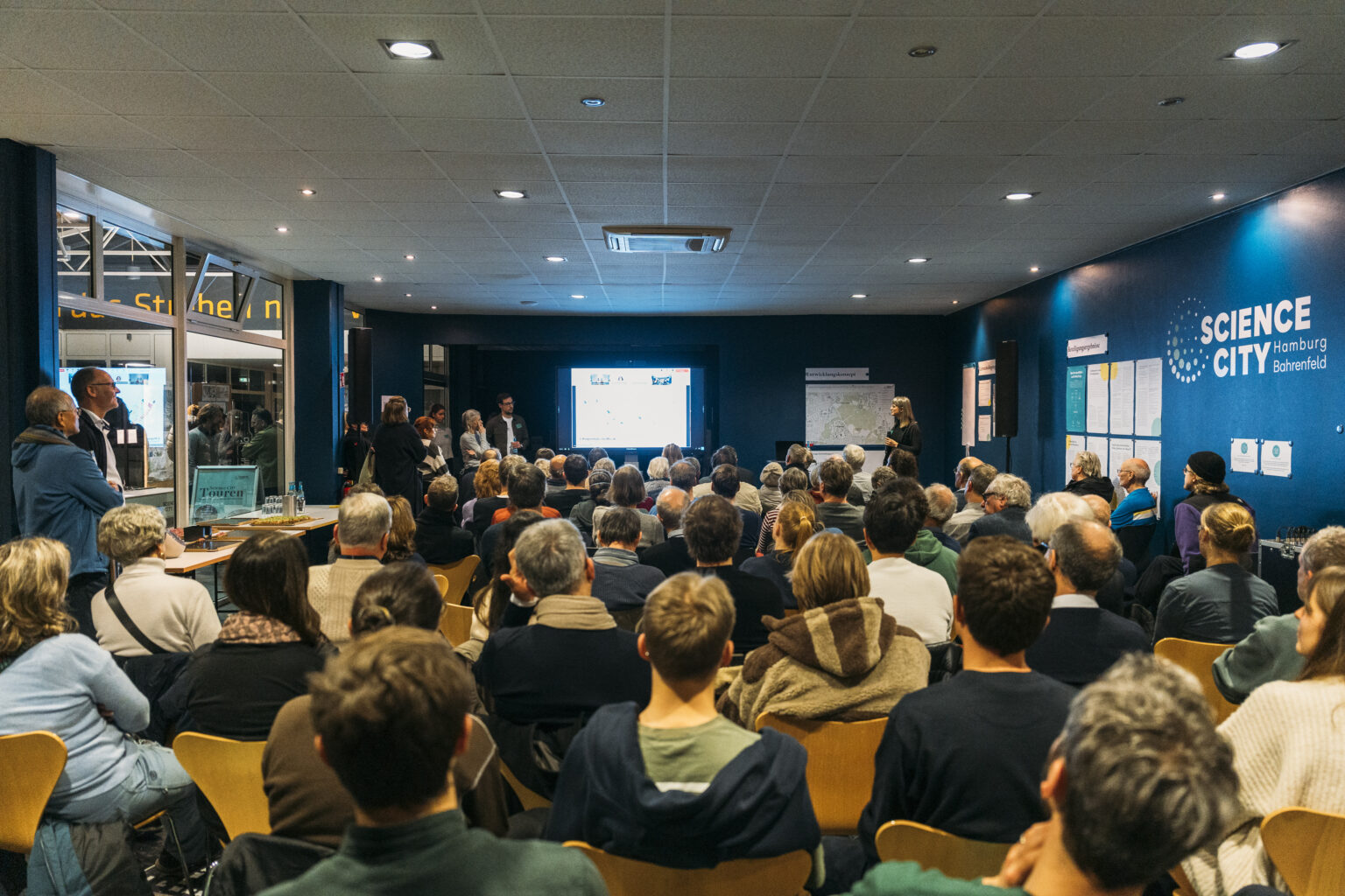 Several people sit in a room while a woman gives a presentation at an event.
